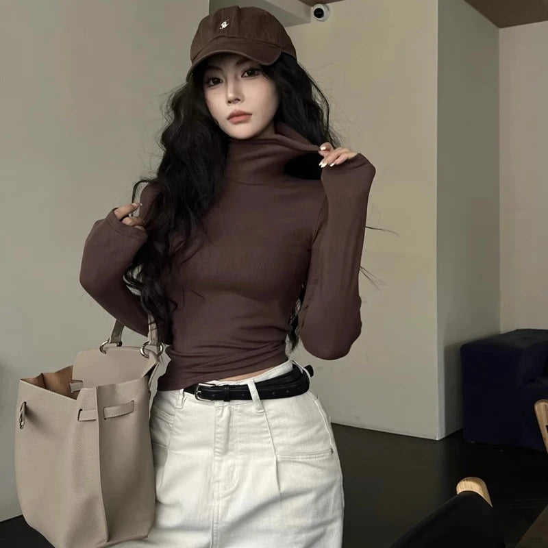 Woman wearing a brown top and white pants, holding a beige handbag indoors.
